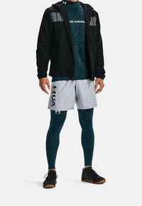 Pánský sportovní outfit, který zahrnuje černou zipovou bundu, těsně přiléhající tealovou košili, šedé šortky v camo vzoru a černé tréninkové boty. Viditelné detaily texturované látky.