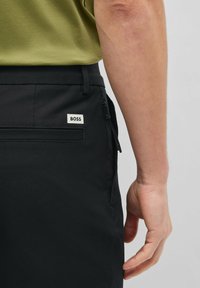 Black trousers with side zip pocket, featuring a fabric tag labeled "BOSS." Smooth, matte texture with a tailored fit.
