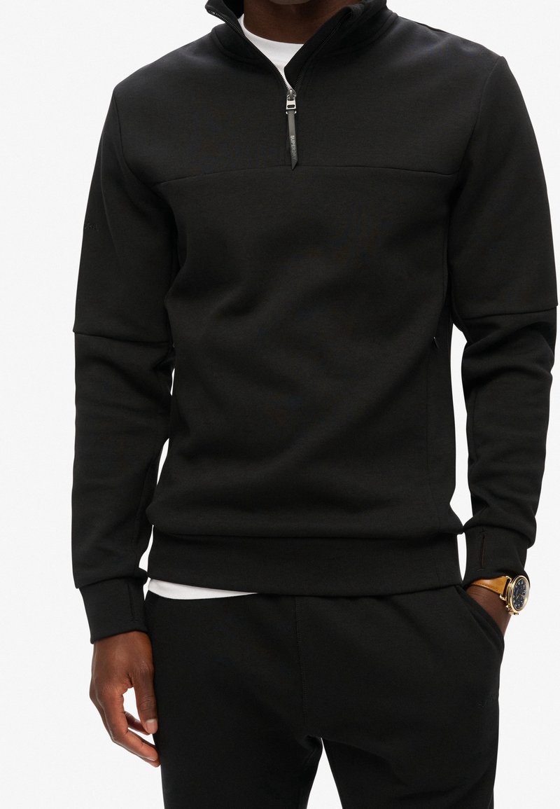 Homme portant un sweat noir à fermeture éclair quart, un pantalon noir, un t-shirt blanc en dessous et une montre-bracelet dorée, debout avec une main dans la poche.