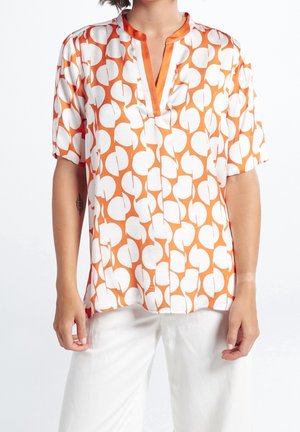 Femme portant un chemisier à manches courtes à motifs géométriques orange et blanc avec col en V et pantalon blanc, vue des épaules aux hanches.