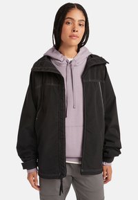 Chaqueta exterior negra con un ajuste holgado, que presenta costuras blancas en contraste y una capucha. Lleva una sudadera ligera de color púrpura debajo. Textura de tela suave.