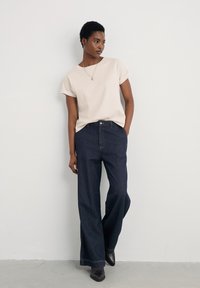 Top beige a maniche corte abbinato a jeans a gamba larga blu scuro. I jeans hanno una chiusura classica con bottone e cuciture visibili lungo l'orlo.