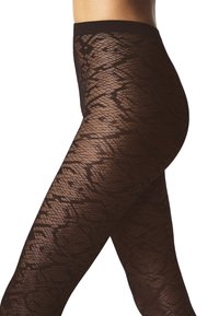 Gambe incrociate, indossando collant trasparenti neri con motivo pitone, visibili dalla vita ai piedi su uno sfondo bianco.