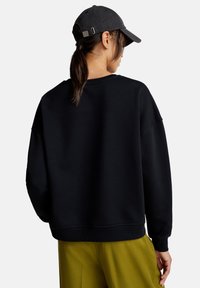 Felpa nera con scollatura tonda e spalle basse, abbinata a un cappellino grigio e pantaloni verdi oliva, vista da dietro.