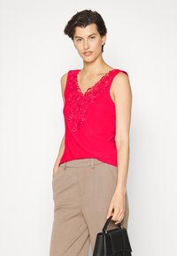 Top vermelho sem mangas com decote em V, apresentando um intricado bordado floral. Combinado com calças bege e uma bolsa preta.