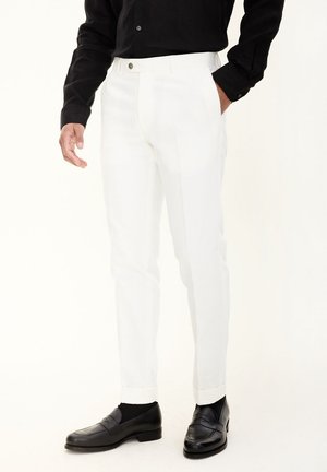 Pantalons blancs ajustés avec une coupe slim et des passants de ceinture, présentant une fermeture éclair à l'avant et une fermeture à bouton. Associés à des mocassins noirs et une chemise à manches longues.