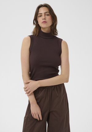 Femme debout, bras croisés, portant un haut à col roulé sans manches marron foncé et un pantalon marron ample sur un fond uni.