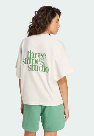 Oversizowa koszulka w kolorze kremowym z zielonym napisem "three stripes studio" na plecach. Połączona z zielonymi szortami, ukazującymi luźny fason.