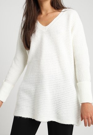 Vrouw draagt een losse, witte gebreide V-hals trui met geribbelde boorden en een zwarte broek tegen een effen achtergrond.