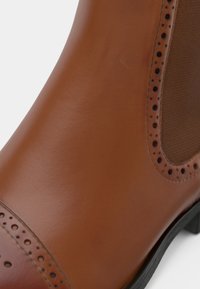 Brown leather ankle boot featuring a smooth finish, brogue perforations, and elastic side panels. Sturdy black sole visible at base.