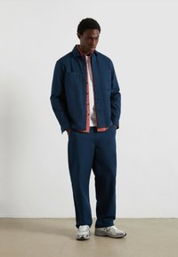 Uomo in piedi con le mani in tasca, indossando una giacca e pantaloni blu navy, una camicia rustica sotto, e scarpe bianche, davanti a una parete bianca uniforme.