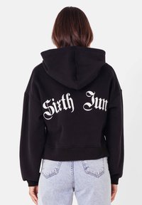 Svart kort hoodie med luva, med vit text "Sixth June" på baksidan. Tillverkad av ett mjukt tyg med långa ärmar.