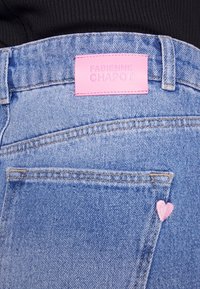 Dettaglio ravvicinato della tasca posteriore di jeans in denim blu con cuori rosa e etichetta del marchio Fabienne Chapot fucsia sulla cintura.