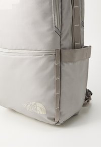 Pelēks mugursoma ar režģa rakstu, aprīkota ar priekšu esošu rāvējslēdzēja kabatu, regulējamām siksnām un logotipa detaļu no The North Face. Izturīga auduma konstrukcija.