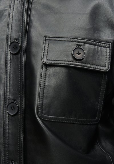 Κοντινό πλάνο μαύρου δερμάτινου μπουφάν που δείχνει την μπροστινή τσέπη με καπάκι που κλείνει με κουμπί και τα διπλανά μαύρα κουμπιά στο εμπρόσθιο μέρος.