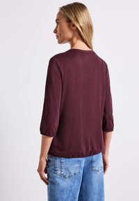 Blouse bordeaux à manches trois-quarts et encolure ronde, dotée d'un ourlet froncé et d'une texture lisse, associée à un jean en denim bleu clair.