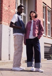To modeller står ute. Den første har på seg en denimvest over en hvit skjorte og beige cargo-bukser. Den andre har på seg en rosa jakke over en hvit topp og mørke jeans. Begge har på seg hvite joggesko.