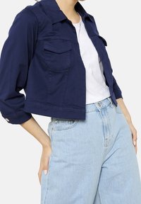 Granatowa, krótka kurtka z kołnierzem, z dwoma kieszeniami na piersi i podwiniętymi rękawami, w zestawieniu z białym T-shirtem i jasnoniebieskimi dżinsami.