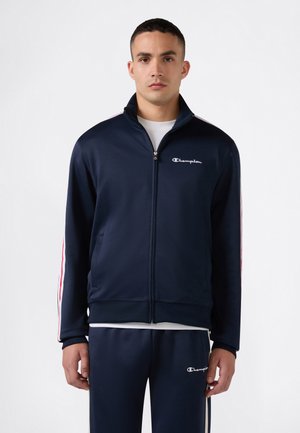 Hombre vestido con un chándal Champion azul marino con rayas blancas y rojas en las mangas y los pantalones, de pie frente a un fondo liso.