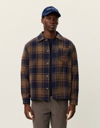 Koszula w kratę w brązowym i granatowym kolorze z zapięciem na guziki, kieszenią na piersi oraz miękką teksturą materiału. Połączona z granatową czapką z napisem.