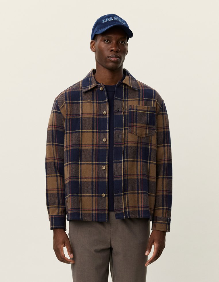 Koszula w kratę w brązowym i granatowym kolorze z zapięciem na guziki, kieszenią na piersi oraz miękką teksturą materiału. Połączona z granatową czapką z napisem.