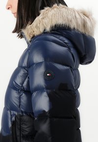 Sieviete, kas valkā tumši zilu pūkaino jaku ar kažokādas apmalēm uz kapuces un Tommy Hilfiger logotipa uzšuvumu augšējā rokā.