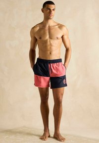 Mężczyzna bez koszuli ubrany w czarne i koralowe kąpielówki w kolorze blokowym, stoi bosy na neutralnym beżowym tle z rękami w kieszeniach.