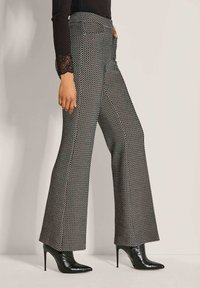 Frau in weit geschnittenen, schwarz-weißen gemusterten Hosen und schwarzen, spitz zulaufenden, hochhackigen Stiefeletten, die vor einem schlichten Hintergrund steht.