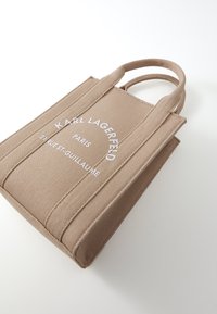 Lett canvas bærepose i lys beige med to håndtak, med hvit tekst: "KARL LAGERFELD PARIS 21 RUE ST-GUILLAUME." Strukturert form med sømdetaljer.