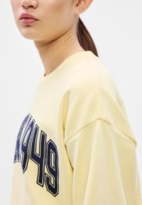 Sárga pulóver tengerészkék "1949." felirattal. Puha anyagból készült, laza szabással és lelógó vállakkal. Simogató tapintású és egyszerű kialakítású.