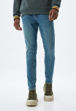 Niebieskie dżinsy o slim fit, z klasycznym pięciopunktowym designem, połączone z zielonymi sneakersami typu high-top oraz szarym sweatshirtem.
