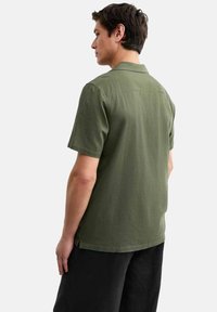 Chemise à manches courtes, couleur olive, en tissu texturé, avec un col montant et un détail d'empiècement au dos, assortie à un pantalon noir.