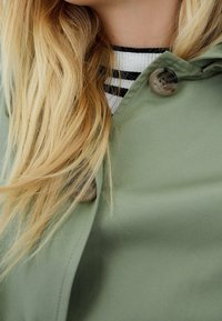 Light green raincoat featuring a two-button front and a smooth texture; layered over a striped black-and-white sweater.