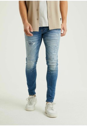 Uomo che indossa jeans skinny azzurri strappati, sneakers bianche, camicia beige a maniche corte con bottoni e maglietta bianca sotto.