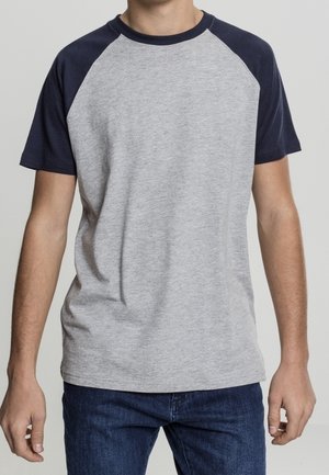 Mand iført en kortærmet raglan T-shirt med grå krop og mørkeblå ærmer, kombineret med blå jeans, stående foran en ensfarvet baggrund.