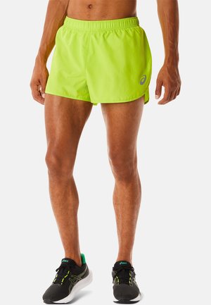 Męski model ubrany w neonowo żółte spodenki do biegania i czarne buty do biegania na jednolitym białym tle.