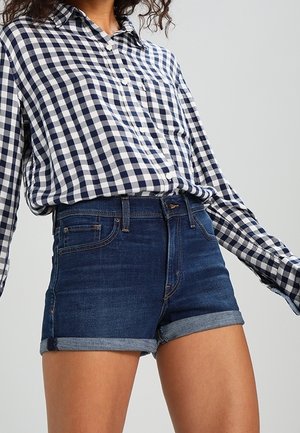 Kvinde iført en marineblå og hvid ternet skjorte med knapper, stoppet ned i mørkeblå denimshorts med opbukket kant på en ensfarvet baggrund.