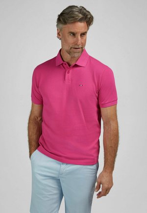 Mężczyzna o siwych włosach, ubrany w jaskraworóżową koszulkę polo i jasnoniebieskie szorty, stoi z jedną ręką w kieszeni, patrząc w bok.