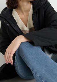 Giubbotto puffo nero con zip frontale, dotato di cappuccio e tessuto lucido. Abbinato a un maglione a righe crema e marrone e jeans blu.