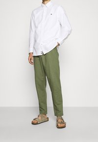 Mand iført hvid langærmet skjorte med knapper, olivengrønne bukser og beige sandals med dobbelt rem, stående med den ene hånd i lommen.