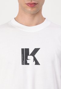 Valkoinen T-paita mustalla "IK"-logolla, jossa on siluetti henkilöstä, jolla on aurinkolasit ja poninhäntä, keskellä rintaa.