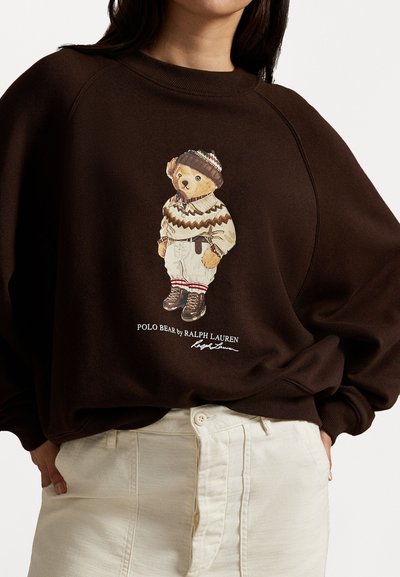 Brūns sporta krekls mīkstā audumā ar teddy bear grafiku ziemas apģērbā, ar svītrām un logo zem tā. Krēmkrāsas bikses redzamas apakšā.