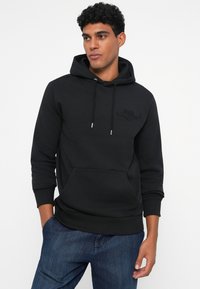 Sudadera con capucha negra, hecha de una tela suave, que presenta un bolsillo frontal tipo canguro y un logo grabado en el pecho. Puños y dobladillo acanalados.