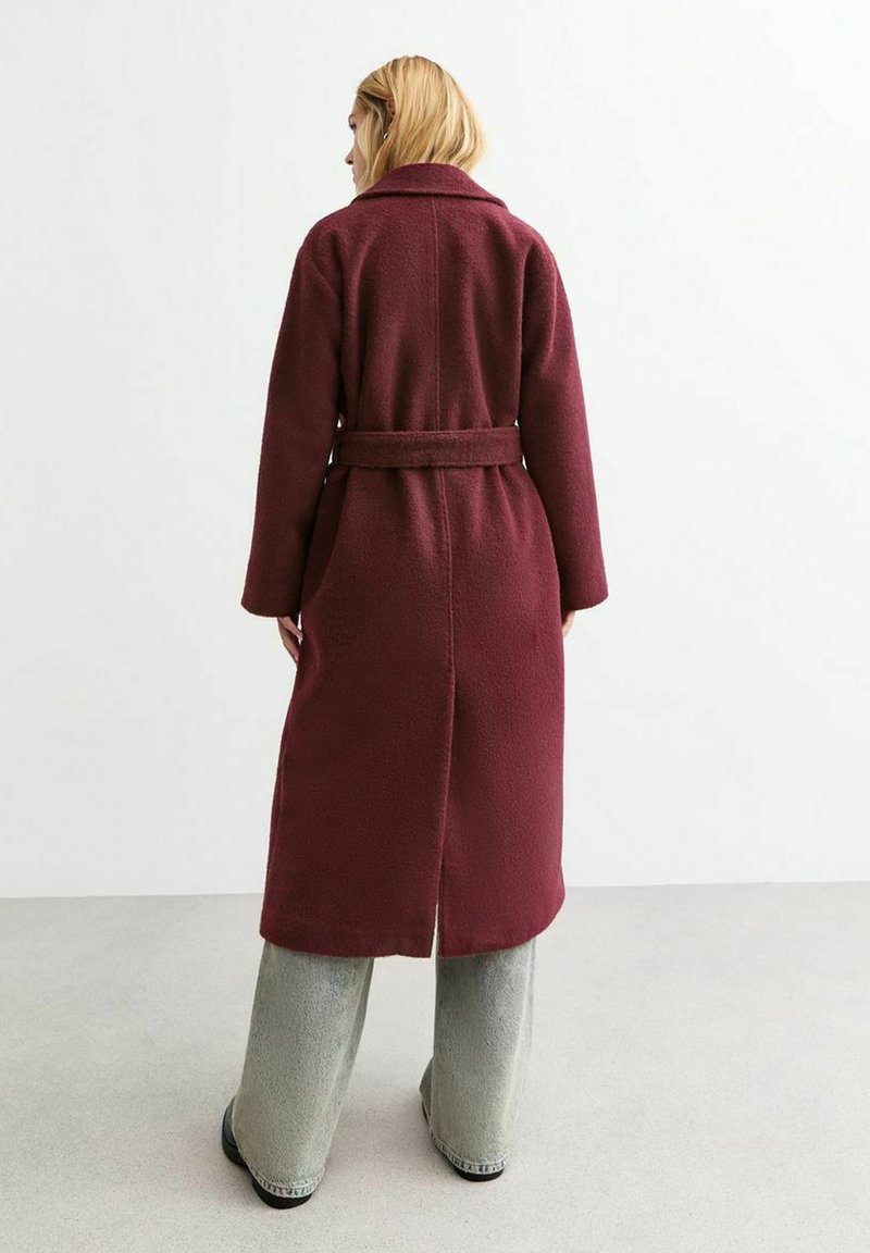 Lange bordeauxrode wollen jas met een riem om de taille, kraag en zijzakken; heeft een split in de rug en wordt gedragen over lichtgrijze wijde broekspijpen.