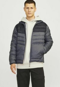 Abrigo acolchado en azul marino oscuro y negro, con cierre de cremallera en la parte delantera, acolchado horizontal, capucha y mangas con puños, llevado sobre una sudadera ligera.