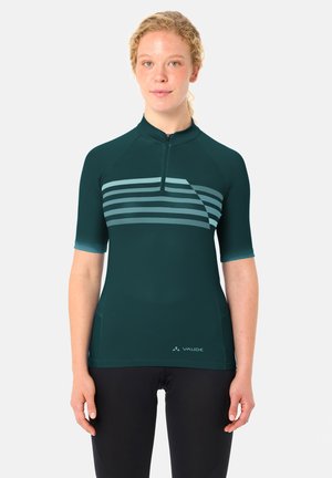 Junge Frau mit lockigem Haar trägt ein dunkelgrünes Radtrikot mit hellblauen horizontalen Streifen und schwarze Radlerhose, steht vor weißem Hintergrund.