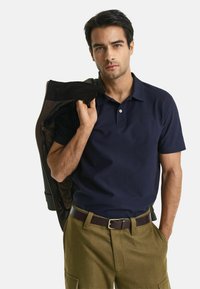 Polo à manches courtes de couleur marine, col plat et deux boutons ; associé à un pantalon vert olive et à une veste foncée portée sur une épaule.