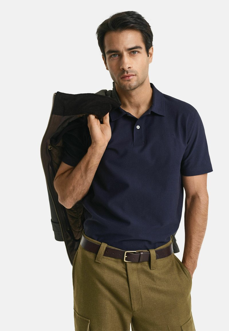 Polo à manches courtes de couleur marine, col plat et deux boutons ; associé à un pantalon vert olive et à une veste foncée portée sur une épaule.