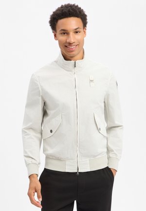 Jeune homme souriant, portant une veste blanche zippée avec poches à boutons et un pantalon noir, debout devant un fond blanc uni.
