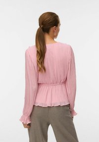 Rosa plisserad blus med långa ärmar, elastisk midja och rynkade manschetter. Bärs med ljusgrå byxor.